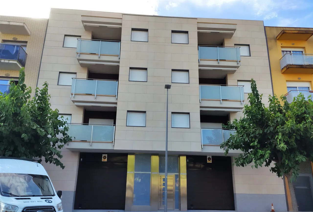 Edificio de viviendas de cuatro plantas con fachada de piezas rectangulares en tono arena y balcones de vidrio.