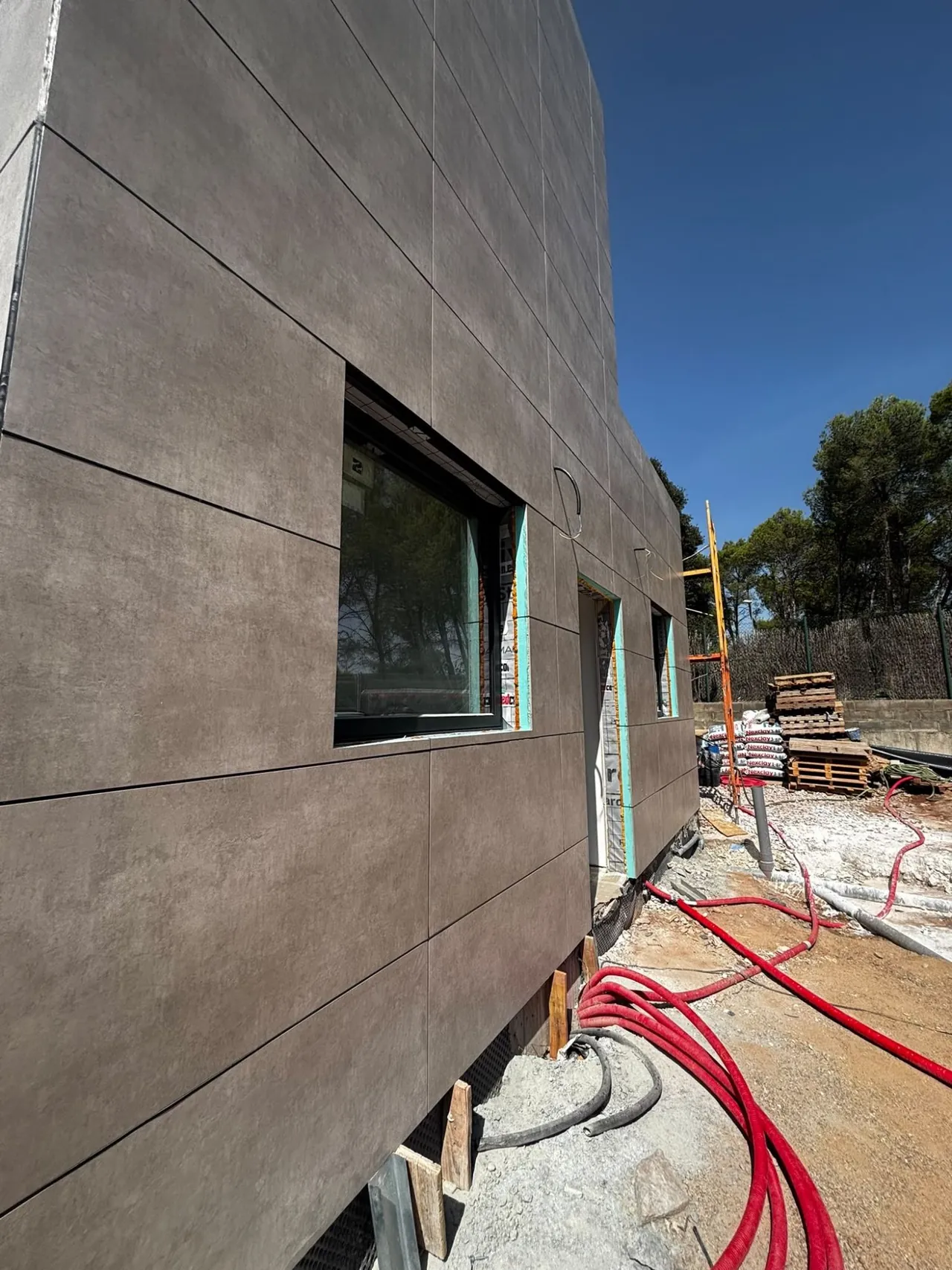 Detalle de una fachada de piezas de gran formato en tono gris pardo, con una ventana, durante la obra.