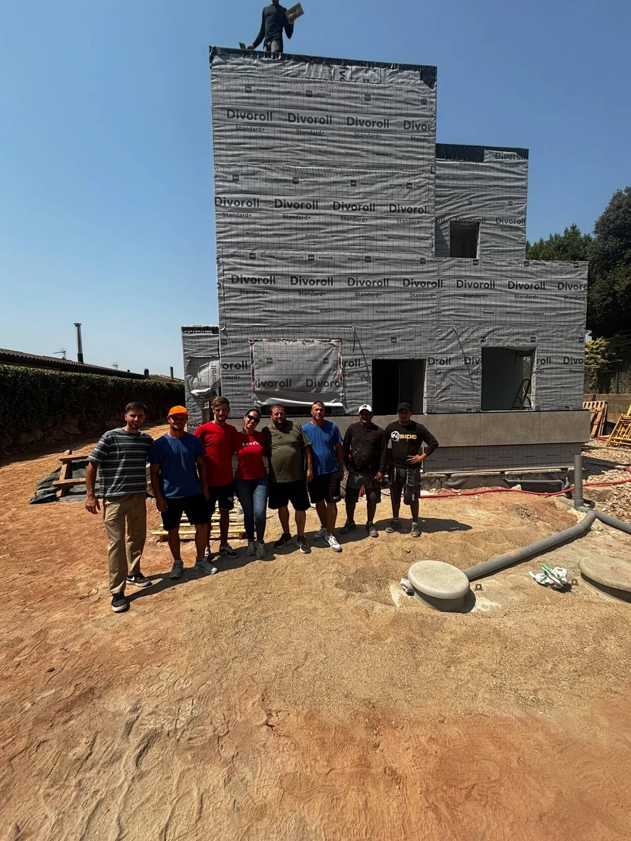 Equipo de obra delante de una vivienda en construcción, con la fachada cubierta por la lámina de protección.